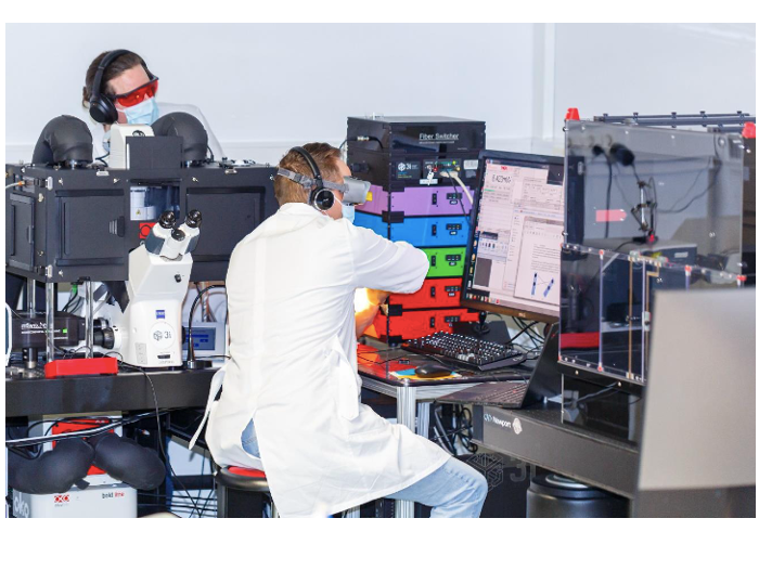 Two white males in lab coats sit in a room surrounded by computer equipment